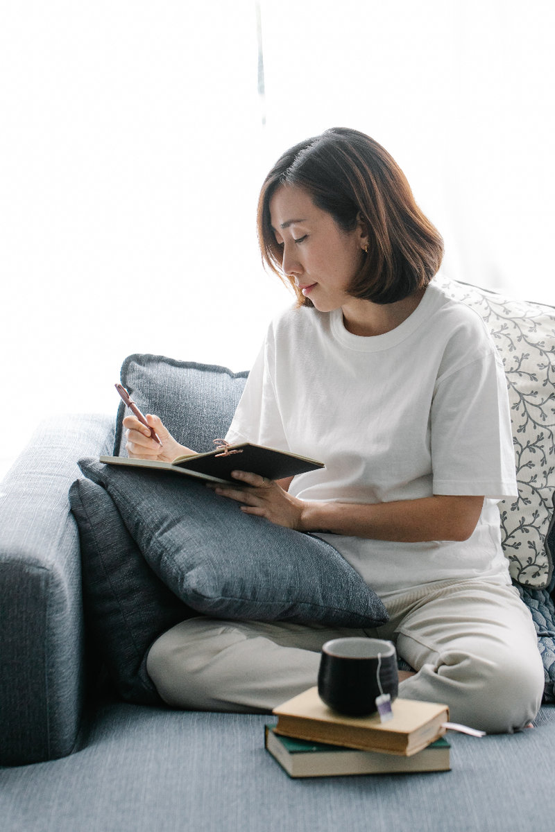 A woman in a white t-shirt and beige pants sits crossed legged on a grey sofa. She has a mug of tea balanced on two hardcover books in front of her, and is using a pillow to prop up and write in her journal.
